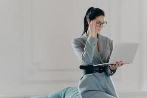 elegante banquera profesional concentrada en la pantalla de la computadora portátil, vestida con ropa formal, lee documentos en poses de versión en línea en una habitación espaciosa, oficina en casa. negocio, concepto de tecnología foto