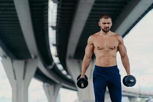 Serious bearded European man with sporty body prepares for barbell workout stands under bridge outdoor has strong biceps and arms concentrated down. Bodybuilding power lifting training concept photo