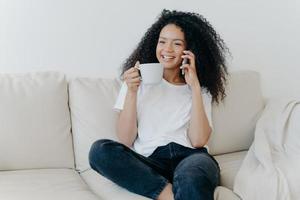 Cheerful Afro American woman has coffee break in living room, sits on couch, calls friend via modern gadget, has happy smile, thoughtful expression, discusses something pleasant. Technology, lifestyle photo