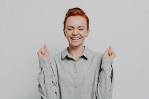 Excited overjoyed young ginger female raising hands with clenched fists, keeping eyes closed photo