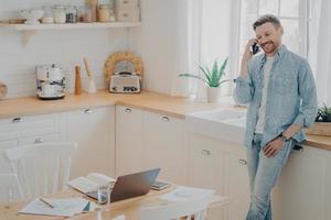 Happy young caucasian man having conversation on cellphone in kitchen photo