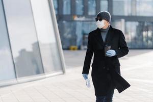 toma horizontal de un hombre adulto serio que lleva una taza de café desechable, vestido con ropa exterior, máscara médica protectora y guantes, se protege durante la pandemia del coronavirus, concentrado a un lado