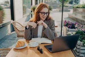 joven mujer sonriente sentada en una mesa de café al aire libre y viendo una lección en línea o un seminario web en una laptop