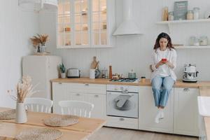 Happy young woman has message on phone. Girl sitting on worktop at kitchen at her apartment photo
