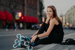 Fit positive woman puts on rollerblades sits on asphalt against blurred street background has positive expression goes in for sport has outdoor fitness activities during summer time. Hobby concept photo