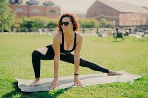 Outdoor workout concept. Atheltic woman stretches on fitness mat goes in for sport regularly poses outside on green lawn looks aside wears black top and leggings has sporty body strong legs. photo