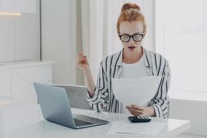 Savings finances concept. Shocked ginger woman focused at document studies bills reads banking paper photo