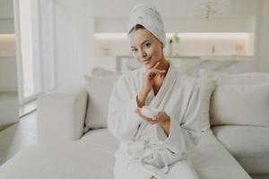 joven mujer hermosa sosteniendo un producto cosmético y sonriendo mientras se relaja en el sofá después de ducharse en casa foto