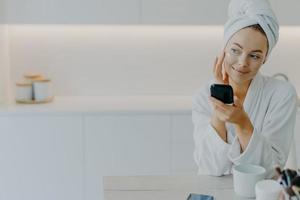 Thoughtful beautiful young woman applies face cream touches cheeks gently holds mirror looks aside dressed in soft bathrobe poses against kitchen interior enjoys cosmetic procedure for skin care photo