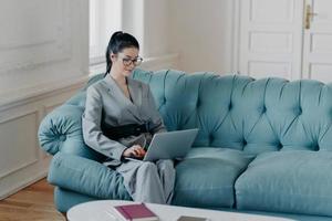 elegante mujer próspera concentrada en la comunicación en línea, ocupada haciendo trabajo a distancia, sentada en un cómodo sofá en un acogedor gabinete, usa una computadora portátil moderna. periodista escribe artículo de publicación foto