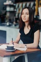 una estudiante pensante escribe una lista para hacer en el bloc de notas, piensa en planes futuros, disfruta de un descanso para tomar café en una cafetería acogedora, anota información con bolígrafo, se concentra en la distancia. gente, concepto de ocio foto