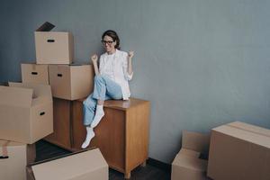 Overjoyed hispanic girl is sitting among cardboard boxes in new house and celebrating. photo