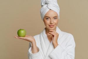 Portrait of good looking young European woman has perfect glowing skin hods fresh green apple undergoes beauty procedures at home dressed in white bath dressing gown isolated over brown background. photo