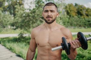 la foto al aire libre de un culturista musculoso con el torso desnudo levanta la barra y tiene un fuerte entrenamiento de bíceps al aire libre mira cuidadosamente a la distancia usa equipos deportivos. atleta guapo con mancuerna