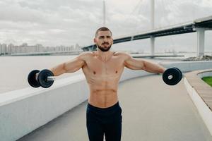 Motivated bodybuilder raises barbells outdoor enjoys great training trains muscles concentrated somewhere poses with naked muscular torso. Athletic shirtless sportsman has workout in open air photo