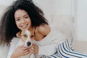 Image of attractive dark skinned woman wears makeup, has bushy curly hair, smiles pleasantly, cuddles dog, dressed in fashionable clothes, enjoys sweet moment with puppy, being at home together photo