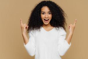 People, advertisement and emotions concept. Glad young female points upwards with index fingers, shows awesome item on sale, wears white sweater, exclaims happily, isolated on brown studio wall photo