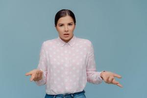 Pretty dark haired woman with makeup, spreads hands and looks doubtfully, feels misunderstanding, wears polka dot shirt and jeans, cannot make decision, poses indoor over blue studio background photo