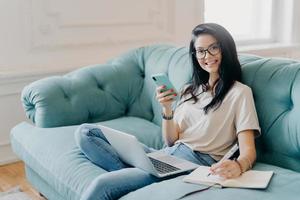 Glad female student works on course paper, surrounded with modern gadget, makes notes in notepad, feels relaxed at comfortable sofa, works online, wears casual clothes, watches video online. photo