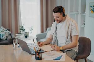 Indoor shot of happy male makes notes while listens online courses studies remotely from home sits at desktop with modern laptop computer papers wears headphones has web conference or virtual meeting photo