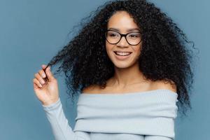 Delighted female holds curl, has bushy hair, being in high spirit, enjoys free time, wears fashionable light blue sweater, shows bare shoulders, stands indoor. People, beauty and happiness concept photo