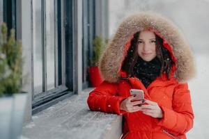 Smiling young European female in red coat, has black manicure, uses modern electronic gadget for communication in internet with friends, likes winter weather, sends photo in social networks.