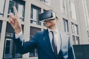 Cheerful businessman trying to touch something in virtual reality, standing near office building photo
