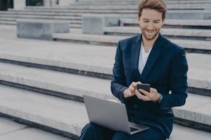 Young nice looking male entrepreneur in stylish suit using smartphone while sits outside with laptop photo