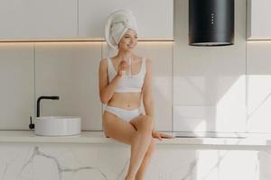 Slim sportive young girl sitting on white marble kitchen table top and drinking glass of mineral pure water photo