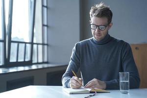 Hombre alemán autónomo seguro de sí mismo con gafas tomando notas en la agenda sentado en el escritorio en casa foto