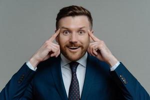 Portrait of handsome male entrepreneur keeps fingers on temples looks with happy surprised expression at camera tries to recall information isolated over grey background. Experienced businessman photo