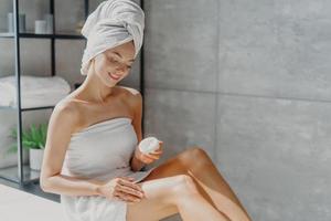 Photo of sensual young European woman uses body cream after taking bath takes care of her skin wrapped in white towel poses against bathroom background. Wellness, pampering and wellbeing concept