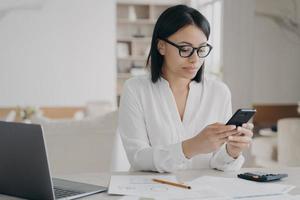 Focused businesswoman in glasses answer client's message using mobile phone apps sitting at laptop photo