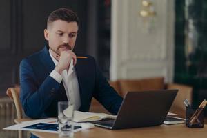 Serious male entrepreneur holds chin makes notes in diary works on laptop computer makes money online dressed formally writes information. Confident managing director involved in working process photo