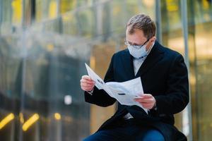 Coronavirus epidemic in city. Horizontal shot of serious man reads newspaper attentively, poses against blurred building background, wears mask for spreading of coronavirus. Businessman with press photo