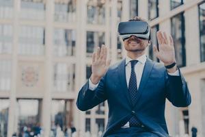 joven trabajador de oficina barbudo feliz vestido formalmente sentado al aire libre y usando auriculares vr o gafas 3d foto