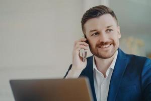 feliz joven apuesto hombre de negocios hablando por teléfono móvil con su pareja frente a la computadora portátil foto