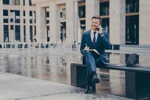 exitoso hombre de negocios hablando por teléfono móvil mientras trabaja afuera, sentado en un banco foto