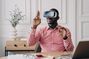 Focused Aframerican man in VR glasses enjoying augmented reality while sitting at workplace photo