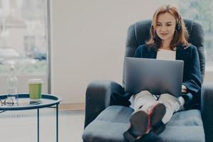 Smiling red-haired woman working in headphones on laptop computer in modern open plan office photo