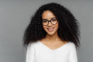 Gorgeous lovely curly woman with Afro hairstyle, feels glad, smiles gently at camera, wears optical glasses and white sweater, isolated on grey background. Happy emotions and feelings concept photo