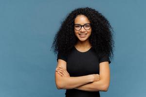 Waist up shot of smiling Afro American woman has arms folded, wears spectacles and casual black t shirt, enjoys nice conversation with interlocutor, poses over blue studio wall with blank space photo