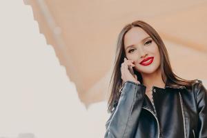 Shot of beautiful young millennial woman enjoys talk with boyfriend, wears leather jacket, has makeup, red lipstick, uses modern cell phone for communication. People and conversation concept photo