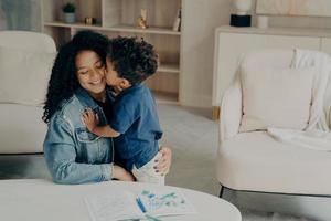 lindo niño pequeño con el pelo rizado abrazando suavemente a su madre sonriente sentada en el suelo en la sala de estar foto