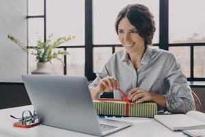 Happy cheerful Italian woman home office worker sits at desk unpacks newly received New Year gift photo