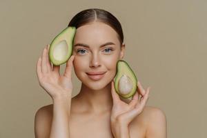 Photo of shirtless female model looks with gentle expression at camera holds halves of avocado near face has healthy well cared skin after cosmetic treatments isolated over beige background.