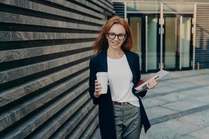 exitosa mujer de negocios de jengibre parada al aire libre cerca de la oficina con laptop, notebook y café foto