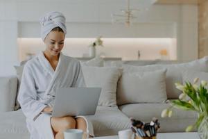Horizontal shot of pretty young woman dressed in bathrobe and towel after shower sits on comfortabe sofa in living room works on laptop computer has own beauty blog types feedback to followers photo