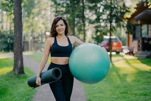 concepto de deporte y recreación. una mujer morena delgada y complacida usa top recortado y polainas, sostiene fitball debajo del brazo, karemat enrollado, posa al aire libre contra el fondo de la naturaleza, se para cerca de los árboles foto