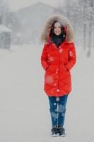 Full length vertical shot of pretty woman wears red winter jacket, jeans and boots, keeps hands in pockets, stands on street during winter time during snowflake, poses for making photo outdoor